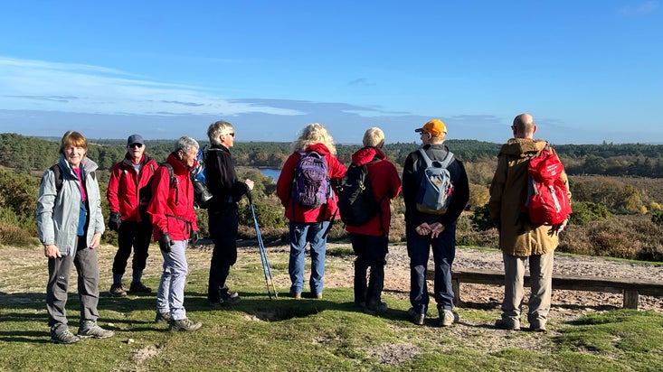 Eight people in walking attire standing at the top of a hill admiring a view. Five people have their backs to the camera and three people are looking at the camera.